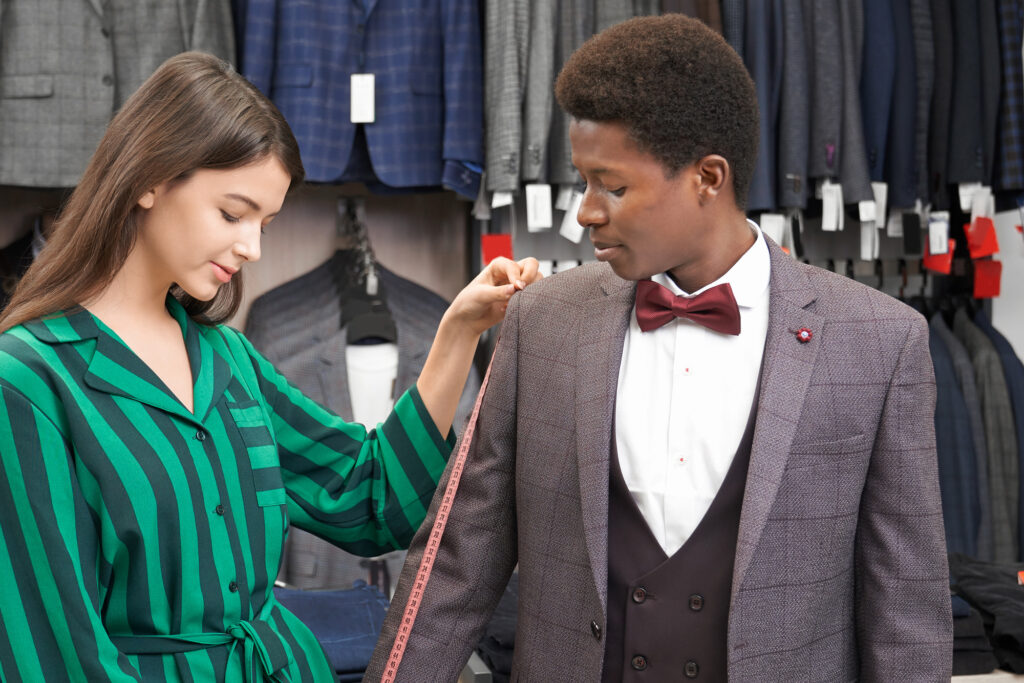 Beautiful female shop consultant choosing suit for young man with curly hair. Brunette holding measuring tape and measuring sleeve of grey jacket on client. Fashion and style.